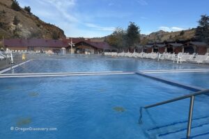 Kah-Nee-Ta Resort and Spa in Oregon: The picture "Kah-Nee-Ta Resort and Spa in Oregon main pool and lodge" shows the large, clear blue outdoor pool in front of the resort’s main lodge building, with white lounge chairs and surrounding hills under a partly cloudy sky.