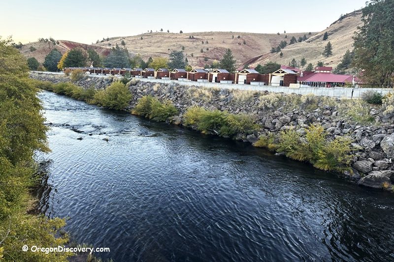 Kah-Nee-Ta Resort and Spa in Oregon: The picture "Kah-Nee-Ta Resort and Spa in Oregon cabins and river view" features a scenic view of a river flowing beside a row of red-roofed cabins, with rolling dry hills and scattered trees in the background.