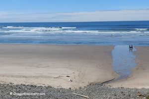 Cove Beach (Falcon Cove Beach aka Magic Rocks Beach) – A small stream of water flows across the sandy beach toward the ocean. Two people sit near the water's edge, while footprints and driftwood are scattered across the sand. The waves stretch out under a partly cloudy sky.