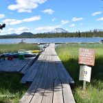 Lava Lake Oregon: A wooden dock extends over the water, passing through tall grass, with boats and canoes moored at the side and a sign listing dock rules; snow-capped mountains and pine trees are visible in the background under a blue sky with scattered clouds.