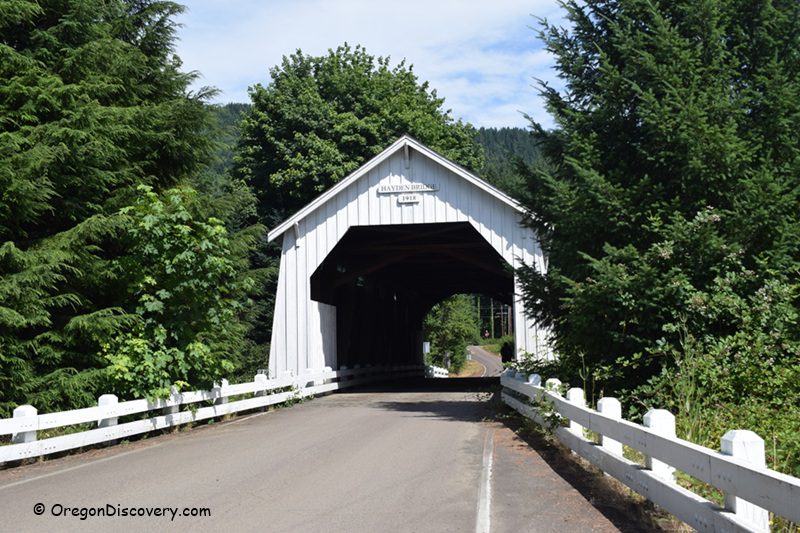 Hayden Covered Bridge Oregon Covered Bridge: A white wooden covered bridge with a peaked roof and white railings, surrounded by lush green trees, with a road leading through it.
