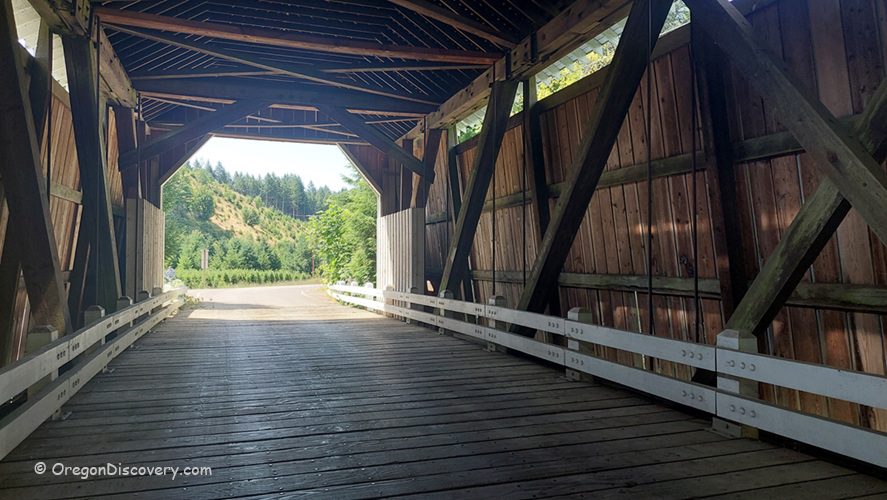 Hayden Covered Bridge Oregon Covered Bridge: Inside view looking out from the wooden bridge, with white railings and beams, opening to a tree-covered hillside and blue sky.
