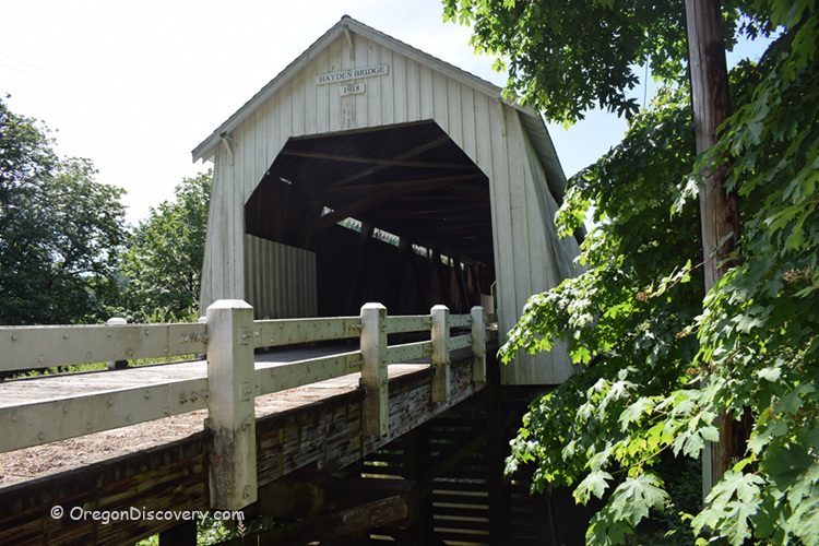 Hayden Covered Bridge Oregon Covered Bridge: Side view of the entrance to the white Hayden Covered Bridge, with wooden guardrails and leafy green trees in the foreground.
