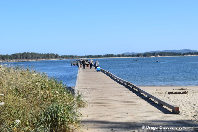 Winchester Bay Oregon: The picture "Winchester Bay Oregon pier" features a long wooden pier stretching over the water with people walking and fishing, surrounded by blue water and grassy shoreline.