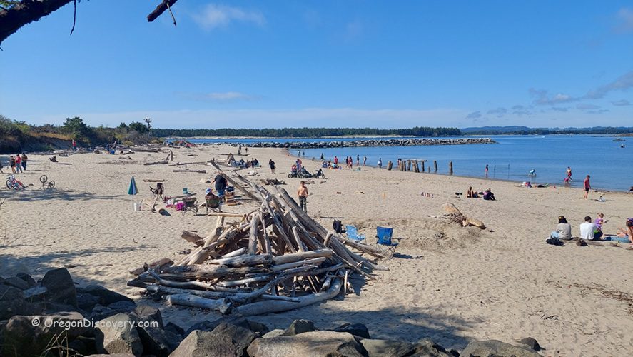 Ziolkouski Beach Park Oregon: Several groups enjoying a sandy beach with scattered driftwood, bicycles, and beach chairs, next to calm blue water and a jetty in the distance.