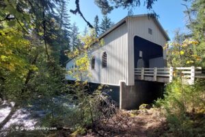 Belknap Covered Bridge in Oregon: The picture "Belknap Covered Bridge in Oregon entrance with road and weight limit sign" features the front entrance of the bridge, painted white, with a paved road leading inside, surrounded by tall trees and a weight limit sign on the right.