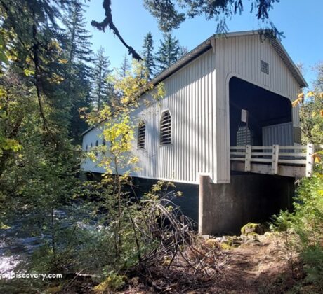 Belknap Covered Bridge in Oregon: The picture "Belknap Covered Bridge in Oregon entrance with road and weight limit sign" features the front entrance of the bridge, painted white, with a paved road leading inside, surrounded by tall trees and a weight limit sign on the right.