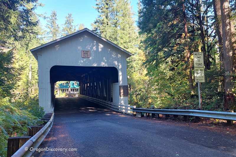 Belknap Covered Bridge in Oregon: The picture "Belknap Covered Bridge in Oregon entrance with road and weight limit sign" features the front entrance of the bridge, painted white, with a paved road leading inside, surrounded by tall trees and a weight limit sign on the right.