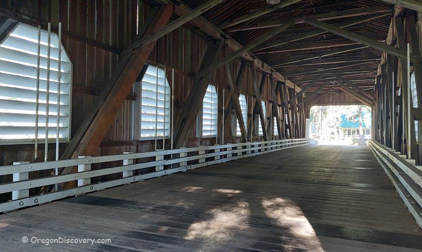 Belknap Covered Bridge in Oregon: The picture "Belknap Covered Bridge in Oregon interior with wooden beams and sunlight" shows the inside of the covered bridge, with wooden beams and white railings along the sides, and sunlight streaming through the louvered windows.