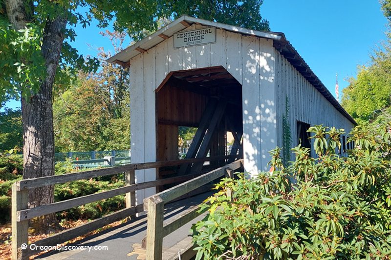 Centennial Covered Bridge in Oregon: The picture "Centennial Covered Bridge in Oregon entrance with ramp" displays the entrance to the covered bridge, with a wooden ramp leading up to the doorway and lush green bushes and trees around the structure.