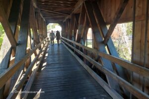 Centennial Covered Bridge in Oregon: The picture "Centennial Covered Bridge in Oregon exterior side view" shows the white wooden bridge with rectangular windows along its side, crossing over a calm creek and surrounded by leafy green trees on a sunny day.