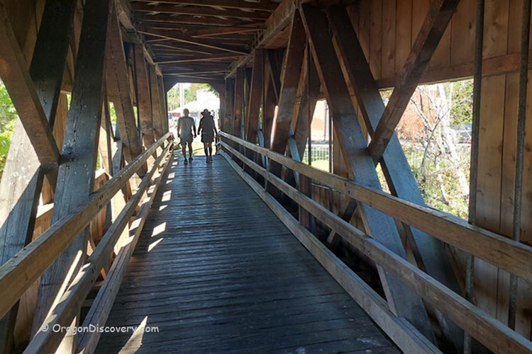 Centennial Covered Bridge in Oregon: The picture "Centennial Covered Bridge in Oregon interior walkway" features the inside view of the bridge, with wooden beams and railings, as two people walk away from the camera toward the far exit.