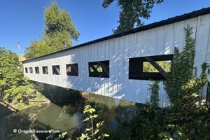 Centennial Covered Bridge in Oregon: The picture "Centennial Covered Bridge in Oregon exterior side view" shows the white wooden bridge with rectangular windows along its side, crossing over a calm creek and surrounded by leafy green trees on a sunny day.