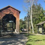 Chambers Covered Railroad Bridge in Oregon: The picture "Chambers Covered Railroad Bridge in Oregon bridge entrance and train sculpture" displays the entrance to the covered bridge with a large metal sculpture of a train engine, nearby park signs, green grass, and trees.