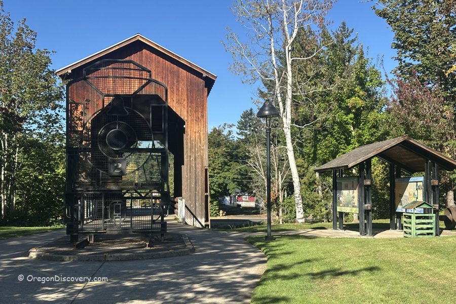 Chambers Covered Railroad Bridge in Oregon: The picture "Chambers Covered Railroad Bridge in Oregon bridge entrance and train sculpture" displays the entrance to the covered bridge with a large metal sculpture of a train engine, nearby park signs, green grass, and trees.