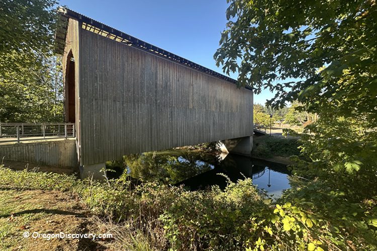 Chambers Covered Railroad Bridge in Oregon: The picture "Chambers Covered Railroad Bridge in Oregon side view with foliage" shows a side view of the covered bridge stretching over the river, framed by green trees and bushes with sunlight filtering through the leaves.
