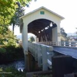 Earnest Covered Bridge in Oregon: The picture "Earnest Covered Bridge in Oregon entrance view over creek" shows the front entrance of the pale yellow covered bridge with white railings, crossing over a creek, surrounded by green trees and sunlight in the background.