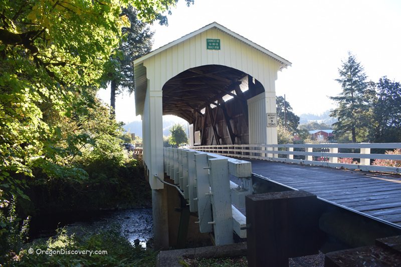 Earnest Covered Bridge in Oregon: The picture "Earnest Covered Bridge in Oregon entrance view over creek" shows the front entrance of the pale yellow covered bridge with white railings, crossing over a creek, surrounded by green trees and sunlight in the background.