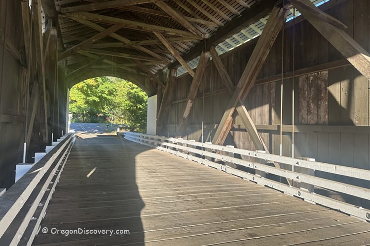 Earnest Covered Bridge in Oregon: The picture "Earnest Covered Bridge in Oregon wooden truss interior" shows the interior of the covered bridge, displaying its wooden truss construction, white railings, and sunlight casting shadows on the wooden floor.