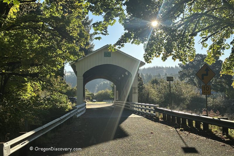 Earnest Covered Bridge in Oregon: The picture "Earnest Covered Bridge in Oregon road approach with sun rays" features the bridge viewed from the road approach, framed by leafy trees with sunlight streaming through the branches, and a road sign visible on the right.