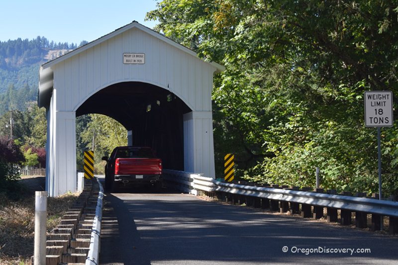 Mosby Creek Covered Bridge in Oregon: The picture "Mosby Creek Covered Bridge in Oregon entrance with red truck" features the white wooden entrance of the bridge, as a red pickup truck drives through, with green trees and mountains in the background and a weight limit sign on the right.