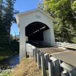 Mosby Creek Covered Bridge in Oregon: The picture "Mosby Creek Covered Bridge in Oregon side view with creek" displays a side view of the white covered bridge stretching over a small creek, surrounded by trees and greenery under a clear blue sky.