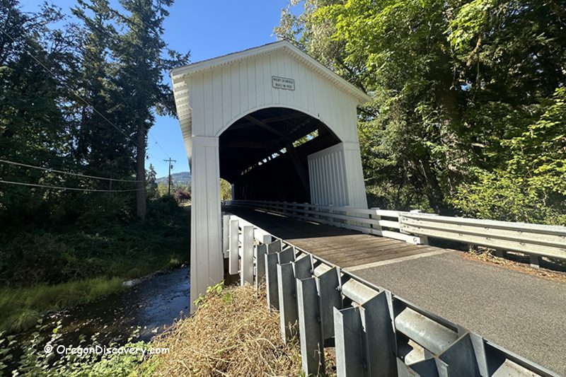 Mosby Creek Covered Bridge in Oregon: The picture "Mosby Creek Covered Bridge in Oregon side view with creek" displays a side view of the white covered bridge stretching over a small creek, surrounded by trees and greenery under a clear blue sky.