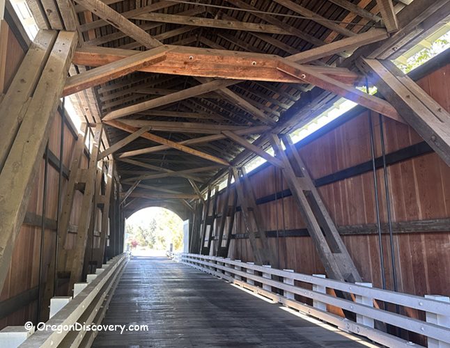 Mosby Creek Covered Bridge in Oregon: The picture "Mosby Creek Covered Bridge in Oregon interior wooden beams" shows the inside of the covered bridge, featuring wooden beams and trusses overhead, with sunlight streaming in from the far entrance.