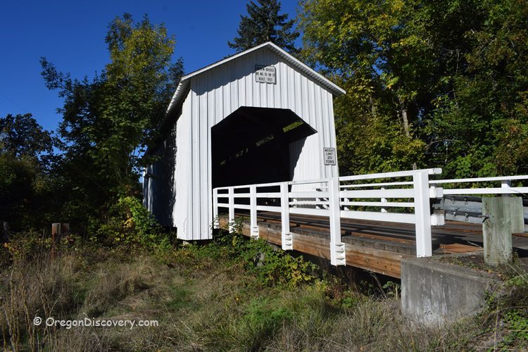 Parvin Covered Bridge in Oregon: The picture "Parvin Covered Bridge in Oregon bridge entrance with white railing" shows the white wooden entrance of the Parvin Covered Bridge, with white railings on both sides and surrounded by green trees and grass under a clear blue sky.