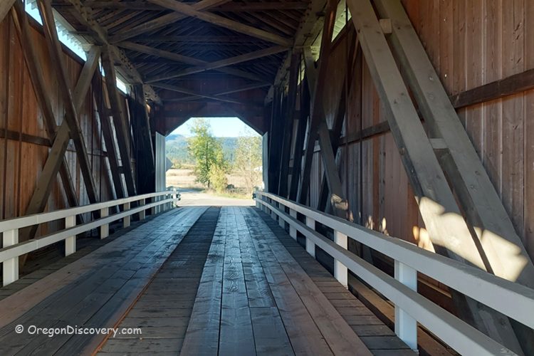 Parvin Covered Bridge in Oregon: The picture "Parvin Covered Bridge in Oregon interior wooden trusses" shows the inside of the covered bridge, with exposed wooden trusses and a plank floor, looking out to the bright landscape at the far end.