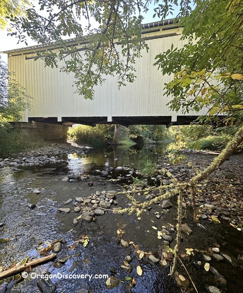 Parvin Covered Bridge in Oregon: The picture "Parvin Covered Bridge in Oregon side view over creek" displays a side view of the white covered bridge crossing a shallow, rocky creek, framed by leafy branches and lush green vegetation.