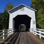 Parvin Covered Bridge in Oregon: The picture "Parvin Covered Bridge in Oregon bridge entrance with white railing" shows the white wooden entrance of the Parvin Covered Bridge, with white railings on both sides and surrounded by green trees and grass under a clear blue sky.