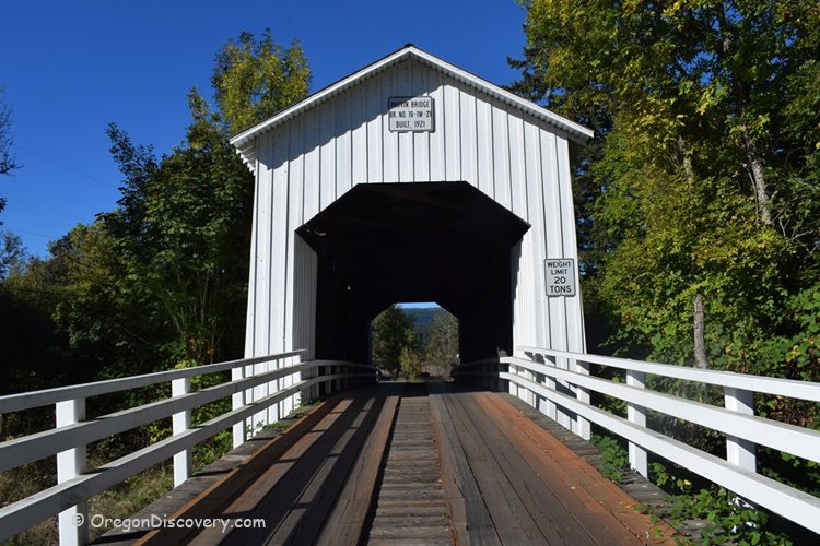 Parvin Covered Bridge in Oregon: The picture "Parvin Covered Bridge in Oregon front view with wooden walkway" features a straight-on view of the bridge’s entrance, highlighting the wooden planks of the road and white railings, with trees and blue sky in the background.