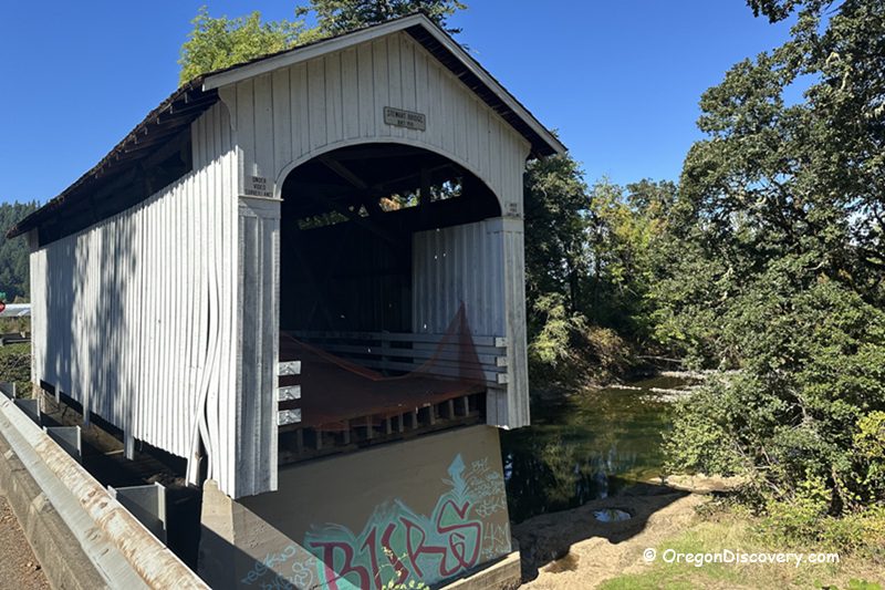 Stewart Covered Bridge in Oregon: The picture "Stewart Covered Bridge in Oregon bridge entrance with graffiti" shows the white wooden entrance of the Stewart Covered Bridge, with some graffiti on the concrete base, looking out over a calm river surrounded by trees.