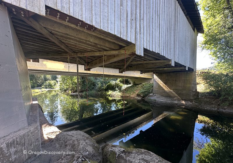 Stewart Covered Bridge in Oregon: The picture "Stewart Covered Bridge in Oregon underside of bridge" shows a close-up view underneath the covered bridge, with its wooden beams and supports reflected in the calm river water below, surrounded by greenery.