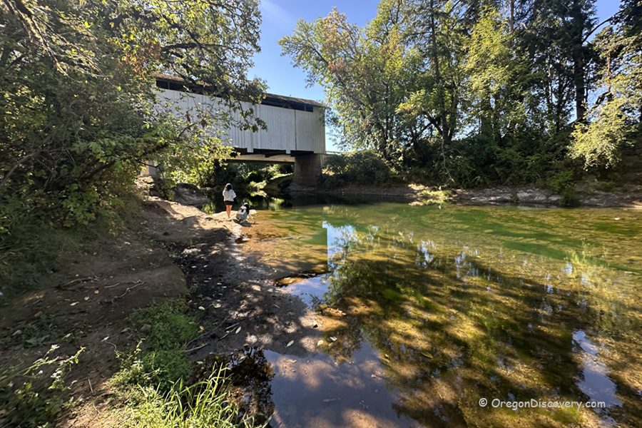 Stewart Covered Bridge in Oregon: The picture "Stewart Covered Bridge in Oregon bridge over river with people" displays a side view of the covered bridge crossing over a shallow, clear river, with two people and a dog standing on the rocky riverbank among leafy trees.