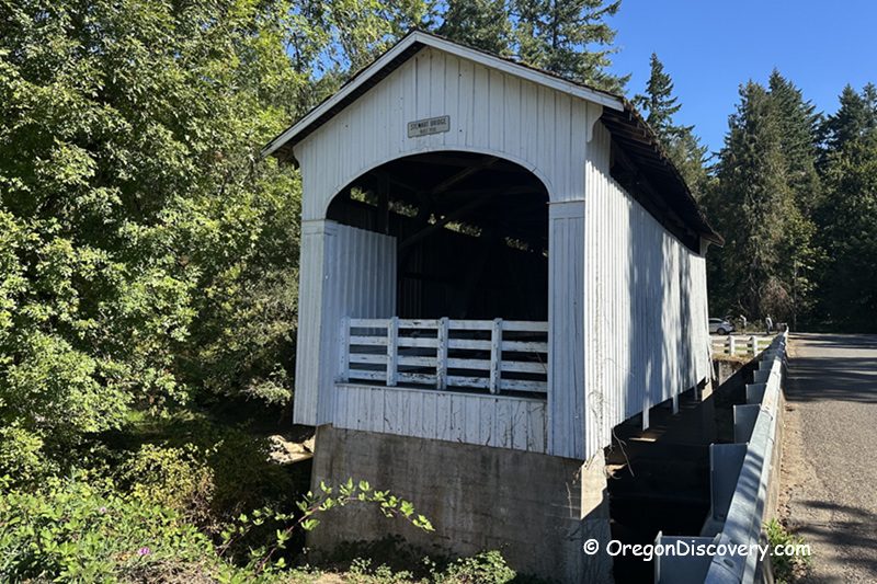 Stewart Covered Bridge in Oregon: The picture "Stewart Covered Bridge in Oregon opposite bridge entrance" features the other end of the white wooden covered bridge, with a view of the road and lush green trees in the background.