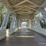 Weddle Covered Bridge in Oregon: The picture "Weddle Covered Bridge in Oregon interior wooden trusses" features the inside of the covered bridge, with white painted wooden trusses and a wooden deck, looking out toward a sunny landscape at the far end.