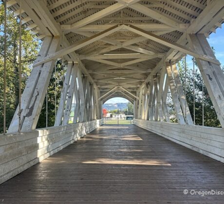 Weddle Covered Bridge in Oregon: The picture "Weddle Covered Bridge in Oregon interior wooden trusses" features the inside of the covered bridge, with white painted wooden trusses and a wooden deck, looking out toward a sunny landscape at the far end.