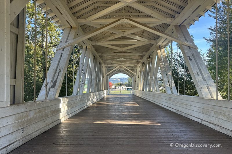 Weddle Covered Bridge in Oregon: The picture "Weddle Covered Bridge in Oregon interior wooden trusses" features the inside of the covered bridge, with white painted wooden trusses and a wooden deck, looking out toward a sunny landscape at the far end.
