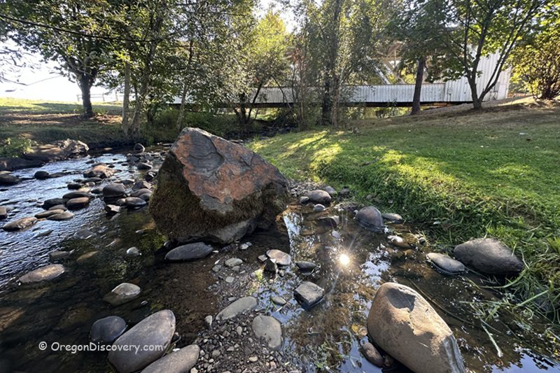Weddle Covered Bridge in Oregon: The picture "Weddle Covered Bridge in Oregon bridge over creek with boulders" displays a side view of the white covered bridge crossing a rocky creek, with a large boulder in the water and grassy banks beneath leafy trees.