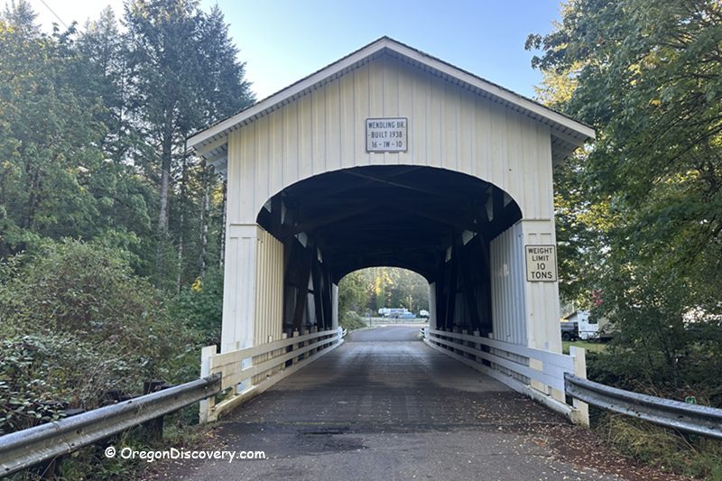 Wendling Covered Bridge in Oregon: The picture "Wendling Covered Bridge in Oregon front entrance view" shows the bridge’s main entrance, with its peaked roof, white siding, a “Wendling Br. Built 1938” sign, white railings, and a road leading through the bridge, framed by trees on both sides.