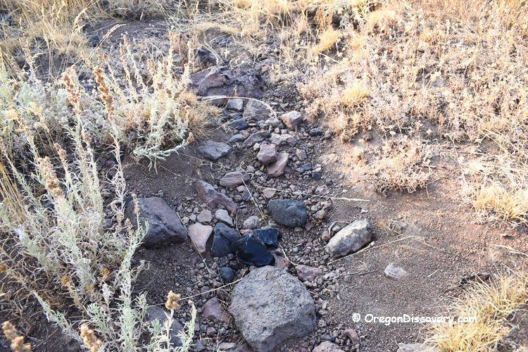 Mud Ridge at Burns - Oregon's Volcanic Glass: The picture "Mud Ridge at Burns black obsidian pieces in dry creek bed" shows a close-up of a shallow, dry creek bed lined with rocks and several shiny black obsidian pieces, surrounded by sparse, dry vegetation.