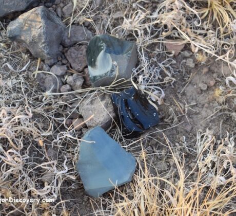Mud Ridge at Burns - Oregon's Volcanic Glass: The picture "Mud Ridge at Burns obsidian and volcanic glass pieces on ground" displays several shiny fragments of obsidian and volcanic glass scattered among rocks and dry grasses on the dusty ground.