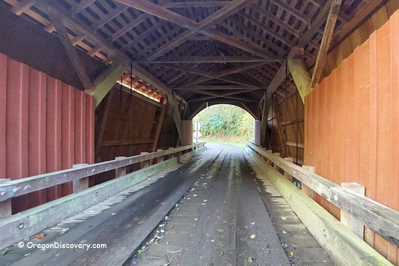 North Fork Yachats Covered Bridge in Oregon: The picture "North Fork Yachats Covered Bridge interior view" shows the inside of the historic wooden bridge, highlighting its exposed beams, red siding, wooden railings, and the road leading through the covered structure toward a leafy landscape.