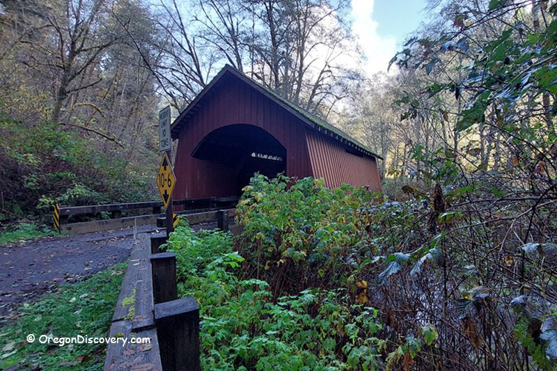 North Fork Yachats Covered Bridge in Oregon: The picture "North Fork Yachats Covered Bridge side angle" features a diagonal view of the red covered bridge surrounded by dense green vegetation and trees, with road signs and a creek visible in the foreground.