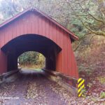 North Fork Yachats Covered Bridge in Oregon: The picture "North Fork Yachats Covered Bridge side angle" features a diagonal view of the red covered bridge surrounded by dense green vegetation and trees, with road signs and a creek visible in the foreground.