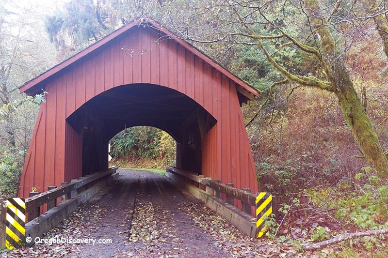 North Fork Yachats Covered Bridge in Oregon: The picture "North Fork Yachats Covered Bridge entrance view" displays the iconic red entrance of the covered bridge, framed by autumn foliage, mossy trees, and wooden railings with yellow and black warning stripes at the base.