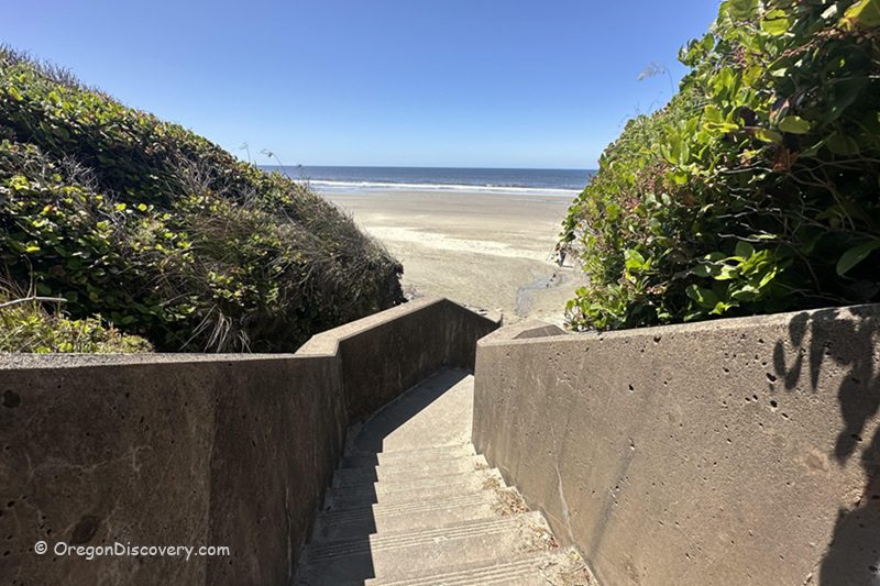 Ocean Beach Picnic Area (Ocean Beach Day Use Site) in Oregon: The picture "Ocean Beach concrete stairs to sand" shows a set of concrete stairs descending between green shrubbery down to a wide sandy beach with the ocean and blue sky in the distance.
