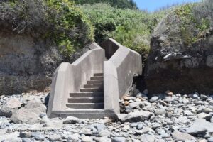 Ocean Beach Picnic Area (Ocean Beach Day Use Site) in Oregon: The picture "Ocean Beach concrete stairs from beach" displays a set of concrete stairs leading up from a rocky beach, positioned between cliffs covered in vegetation.